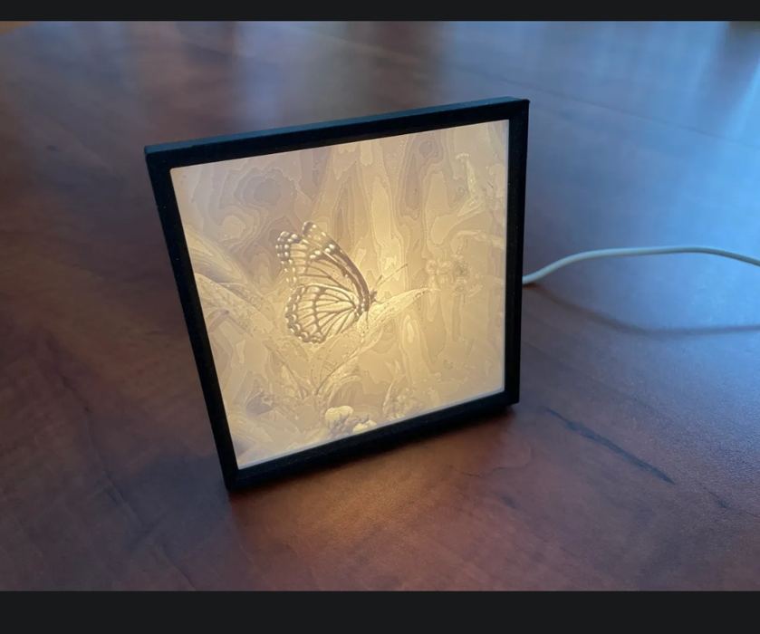 A backlit lithophane frame showing a butterfly on leaves, glowing warm white, with cable